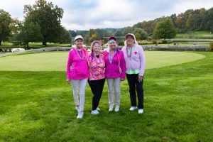 7R9A9941 - Four women pose together on a golf course green, wearing bright pink jackets and hats, with trees and a pond in the background.