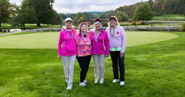 Four women pose together on a golf course green, wearing bright pink jackets and hats, with trees and a pond in the background.
