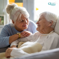 Untitled design - Caregiver with gray hair leaning over a smiling elderly woman in bed, softly holding hands in a warm, comforting moment.