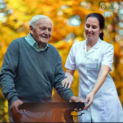 Elderly man using a walker being assisted by a nurse outdoors among orange autumn leaves in a sunlit setting.