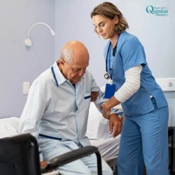 Untitled design 2 - Nurse in blue scrubs with a stethoscope assists an elderly man as he sits on the edge of a hospital bed, using a gait belt for support in a clinical room.