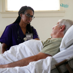 Nurse in purple scrubs with an ID badge and stethoscope speaks to an elderly man resting in a hospital bed.
